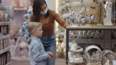 A-mother-in-a-medical-mask-with-her-son-at-the-mall-choose-home-decorations-for-Christmas.-Watch-Christmas-tree-toys.-Coronavirus-epidemic.-Choose-decorations-and-decorations